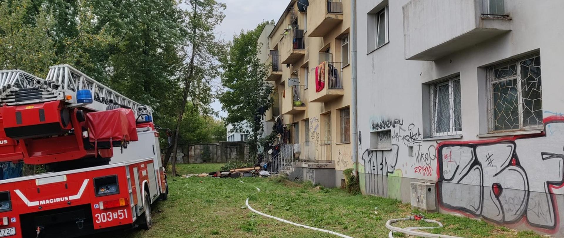 Zdjęcie przedstawia budynek wielorodzinny gdzie doszło do pożaru. Przed nim stoi samochód pożarniczy z drabiną mechaniczną. Na ziemi leżą rozwinięte węże gaśnicze.