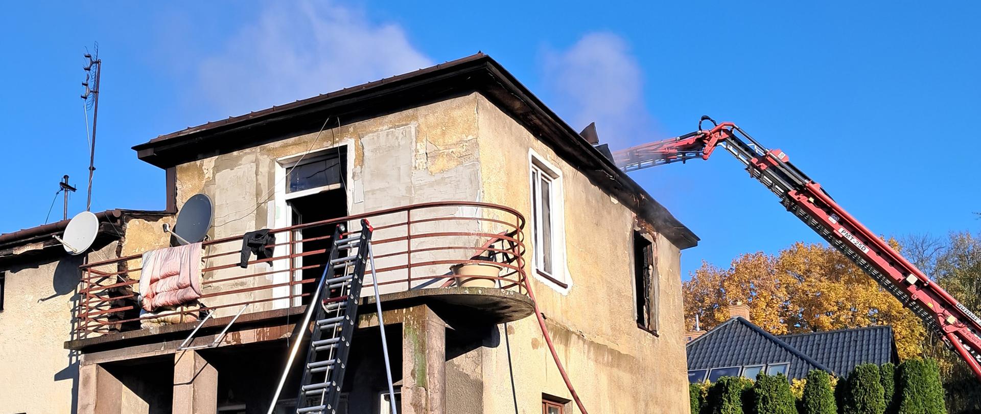 Zdjęcie przedstawia pożar budynku mieszkalnego. Na zdjęciu rozłożone ramie pojazdu pożarniczego drabiny z koszem, który jest nad dachem i prowadzi działania gaśnicze. Z dachu wydobywa się dym. Po lewej stronie do balustrady balkonu przystawiona jest pożarnicza drabina przystawna. Przed budynkiem widać część ogrodzenia.