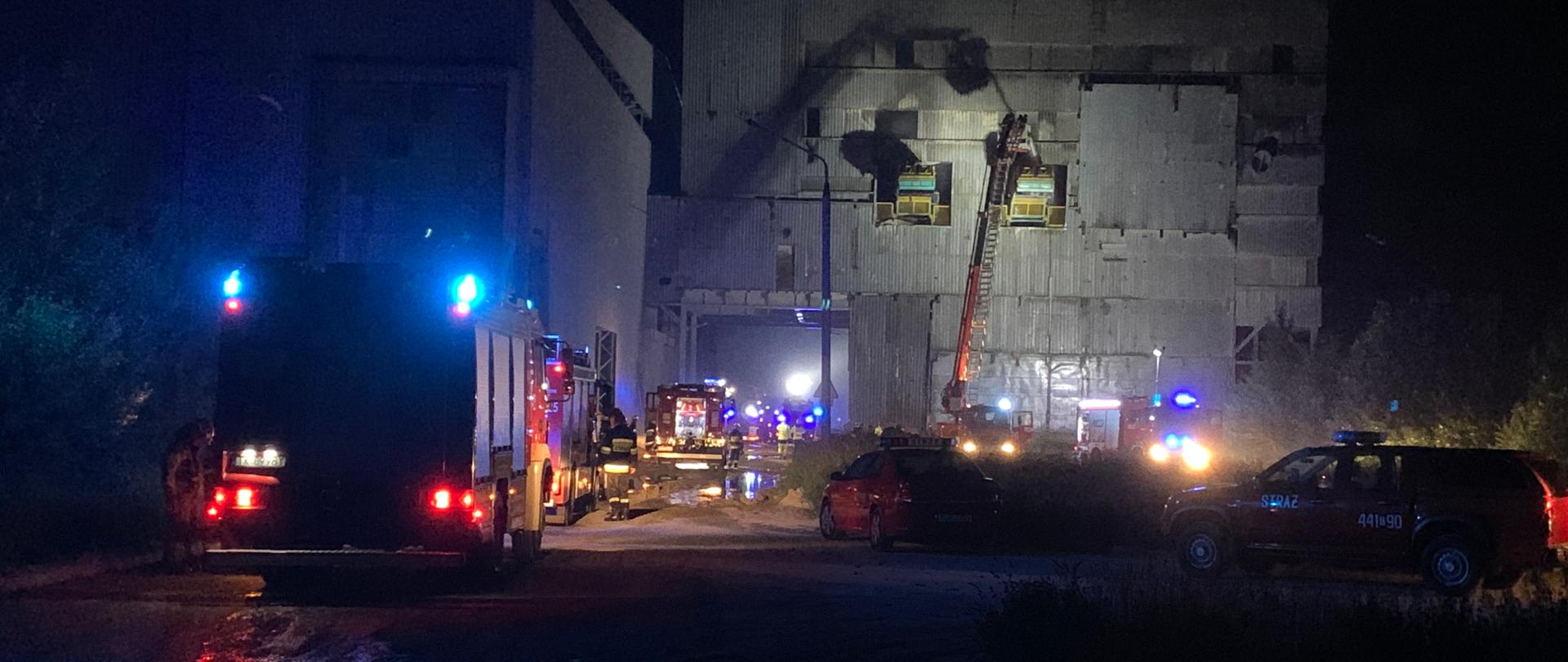 Zdjęcie przedstawia przedstawia pojazdy straży pożarnej z włączonym oświetleniem w nocy, w oddali widoczny podnośnik hydrauliczny przy budynkach cementowni, z którego strażacy gaszą pożar taśmociągów.