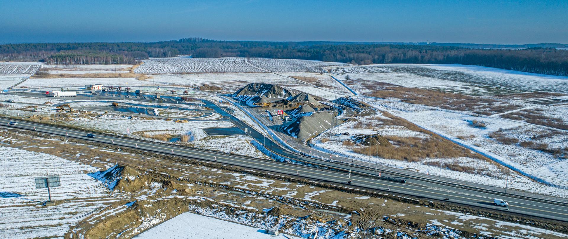 Widok z lotu ptaka na rozbudowywany odcinek drogi ekspresowej. Świeci słońce, zima, widać teren przyprószony śniegiem. Drogą jadą auta, w górnej części zdjęcia widać nasypy, sprzęt budowlany oraz miejsce obsługi podróżnych.