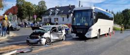 Po prawej stronie stoi autobus koloru białego z rozbitym przodem. Po jego lewej stronie samochód osobowy z rozbitym przodem i drzwiami od strony kierowcy. Obok samochodu stoją strażacy i policjant. W tle 2 domy i gapie. 