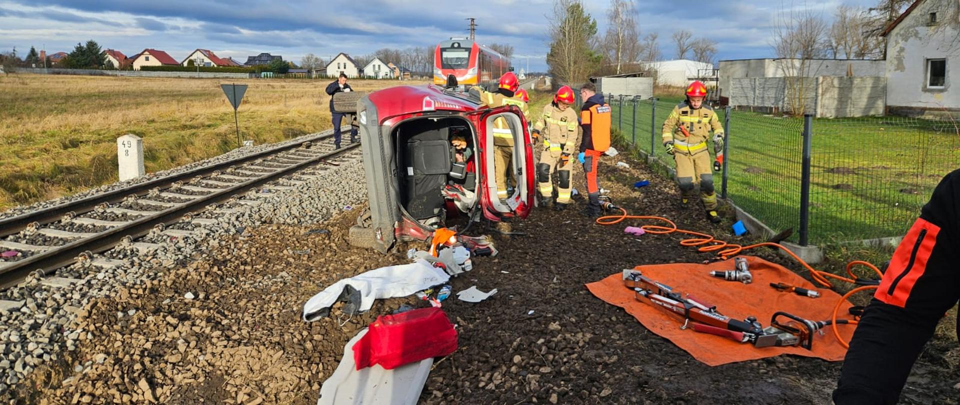 Służby działają w trakcie zdarzenia w wyniku którego samochód osobowy leży na boku. W oddali widać pociąg