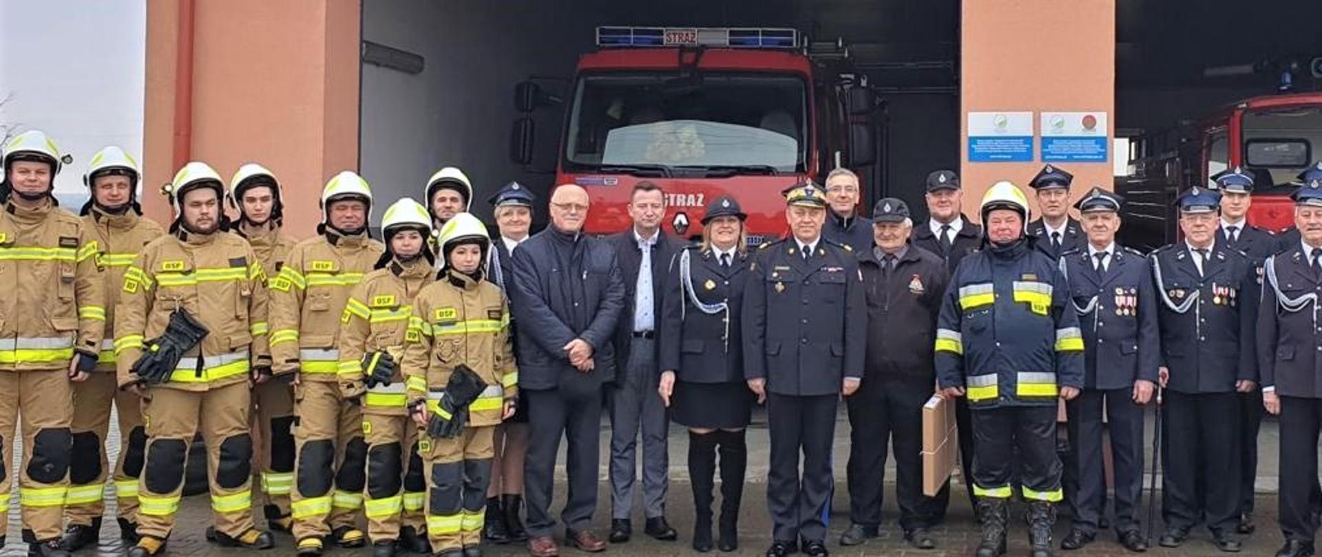 Druhowie Ochotniczej Straży Pożarnej w Niedzborzu stoją przed remizą ustawieni w rzędzie. Na czele stoi nadbrygadier Jarosław Nowosielski Mazowiecki Komendant Wojewódzki Państwowej Straży Pożarnej. Część druhów stojących w rzędzie ubrana jest w stroje specjalne, pozostała część w ubrania galowe.