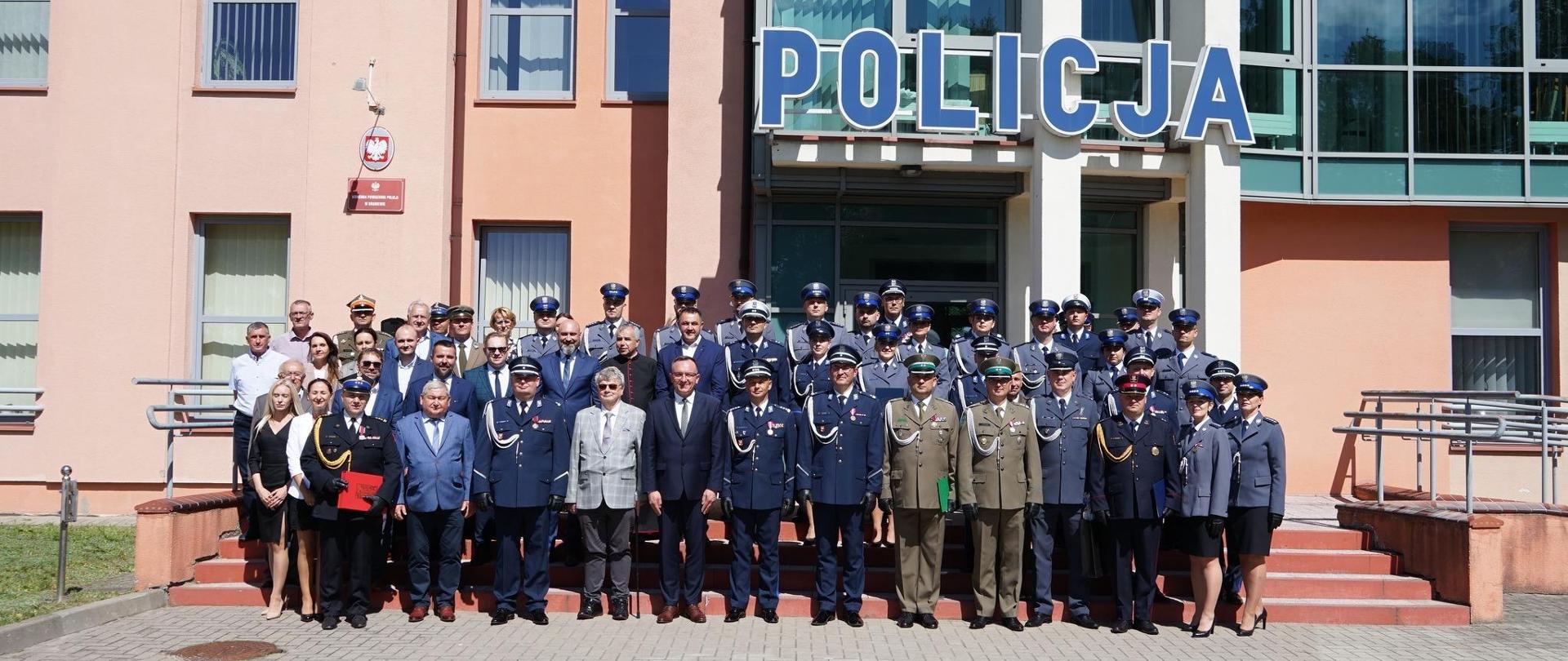 Do zdjęcia pozuje duża grupa funkcjonariuszy i pracowników cywilnych, stoją na schodach przed budynkiem komendy policji. Słonecznie.