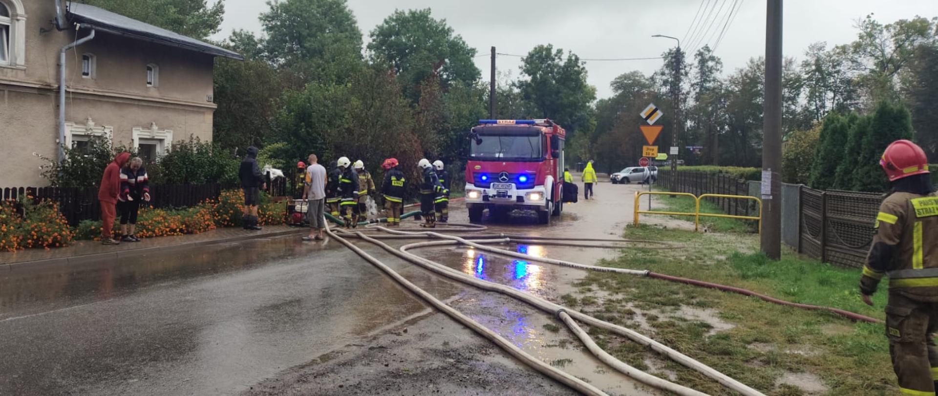 Samochód strażacki z włączonymi światłami, po lewej stronie stoi grupa strażaków, przed pojazdem rozwinięte węże strażackie.