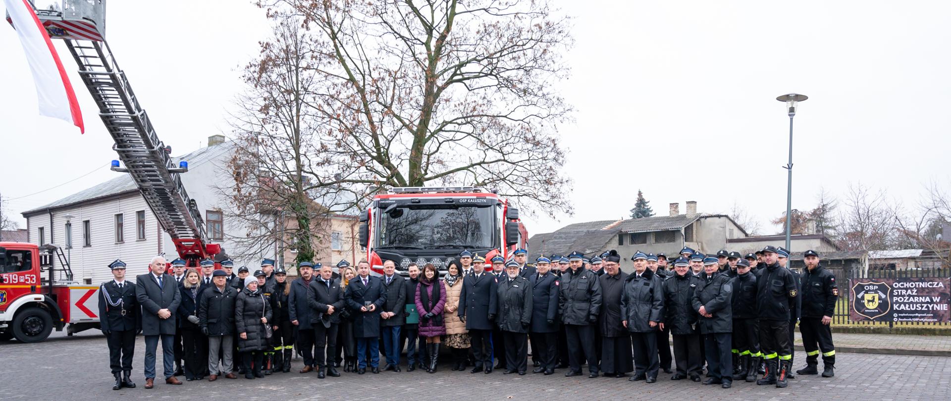 Zdjęcie grupowe uczestników uroczystości na tle samochodu pożarniczego i wysuniętej drabiny drabiny mechanicznej z koszem do którego przymocowana jest biało-czerwona flaga 