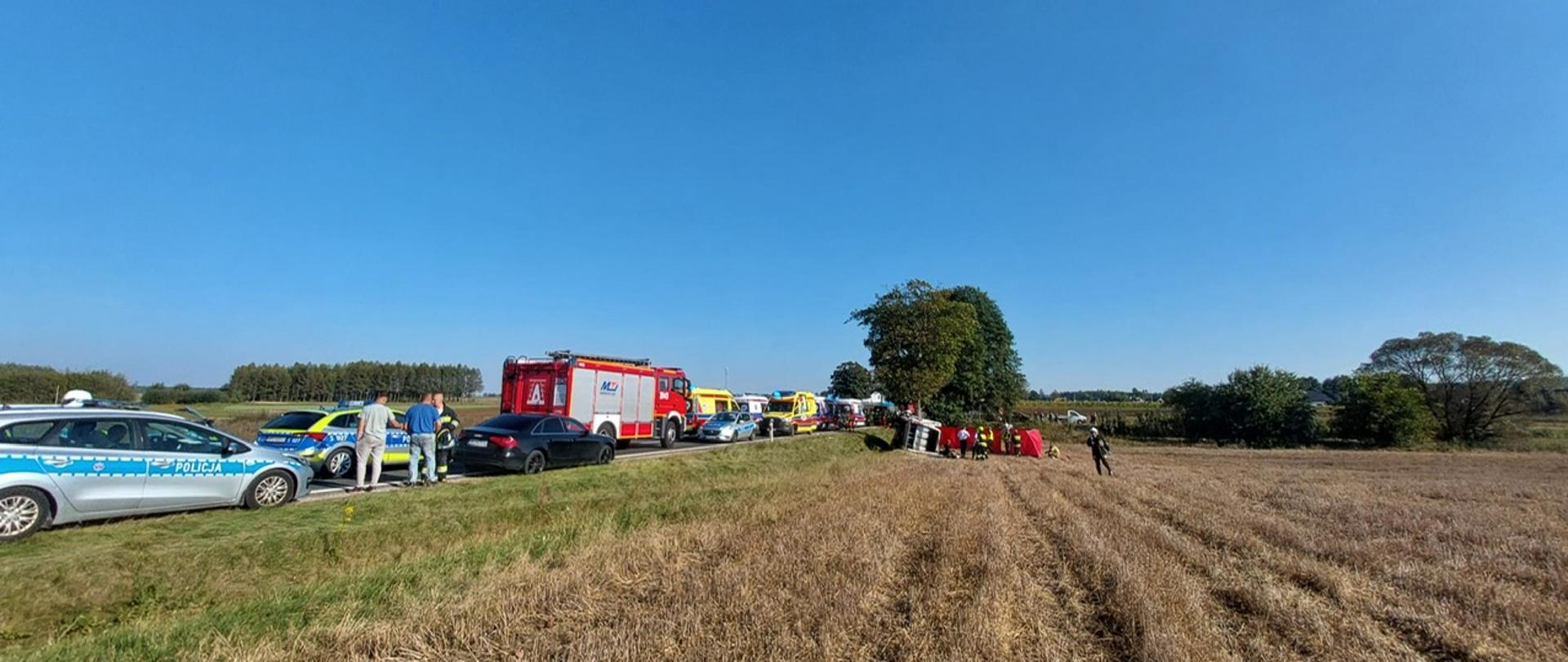 Zdjęcie przedstawia miejsce gdzie doszło do tragedii. Od lewej na drodze stoją 2 radiowozy, wóz strażacki oraz w głębi ambulanse i kolejny radiowóz. W centralnym punkcie zdjęcia widać drzewo, w które uderzył bus. Tam też znajdują się parawany, wrak i skupia się grupa ludzi.