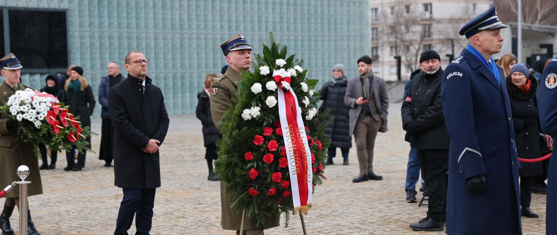 Na zdjęciu mężczyzna niesie wieniec. Za nim idzie przedstawiciel Mazowieckiego Urzędu Wojewódzkiego. 