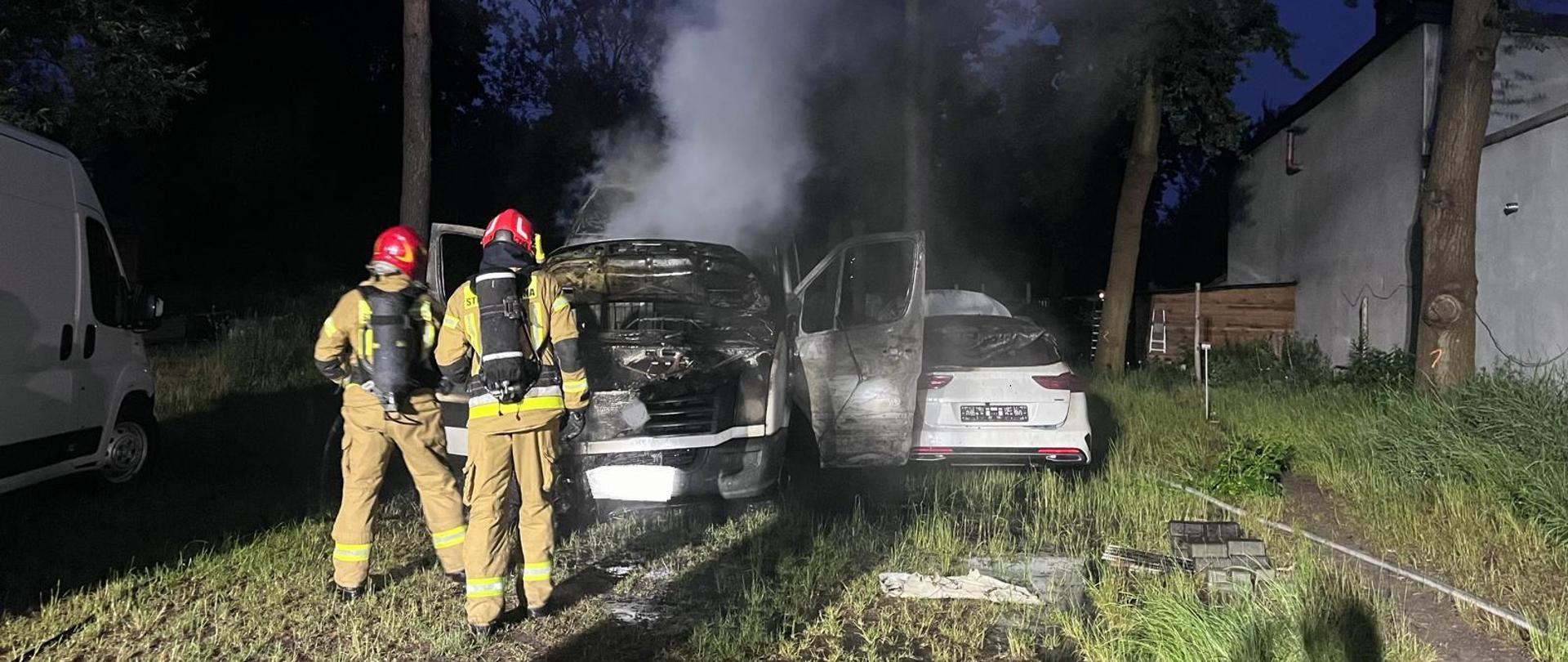 Na zdjęciu widoczne są dwa pojazdy osobowe które uległy spaleniu. Z ich wraków wydobywa się gęsty dym. Przed samochodami stoi dwóch ratowników kontrolujących temperaturę.