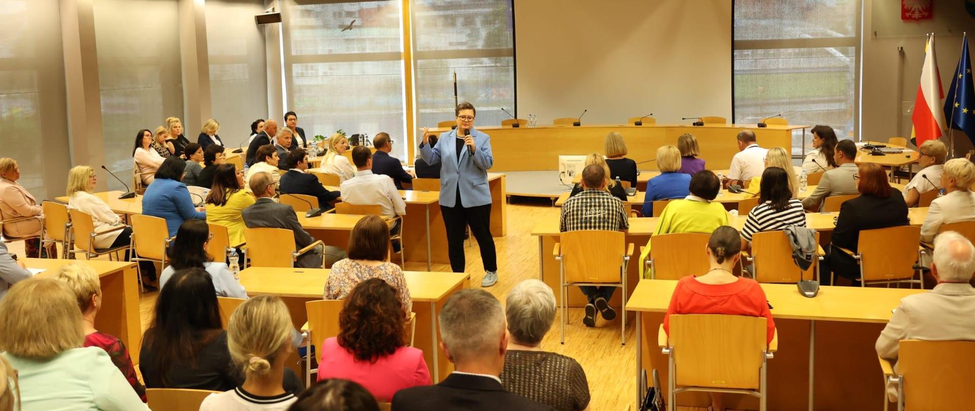 Sekretarz stanu w Ministerstwie Edukacji Narodowej Katarzyna Lubnauer. Na zdjęciu trzyma mikrofon i jest w trakcie przemówienia. Dookoła widzimy pozostałych uczestników spotkania, którzy siedzą przy biurkach w Sali Orła Białego w Urzędzie Marszałkowskim. 