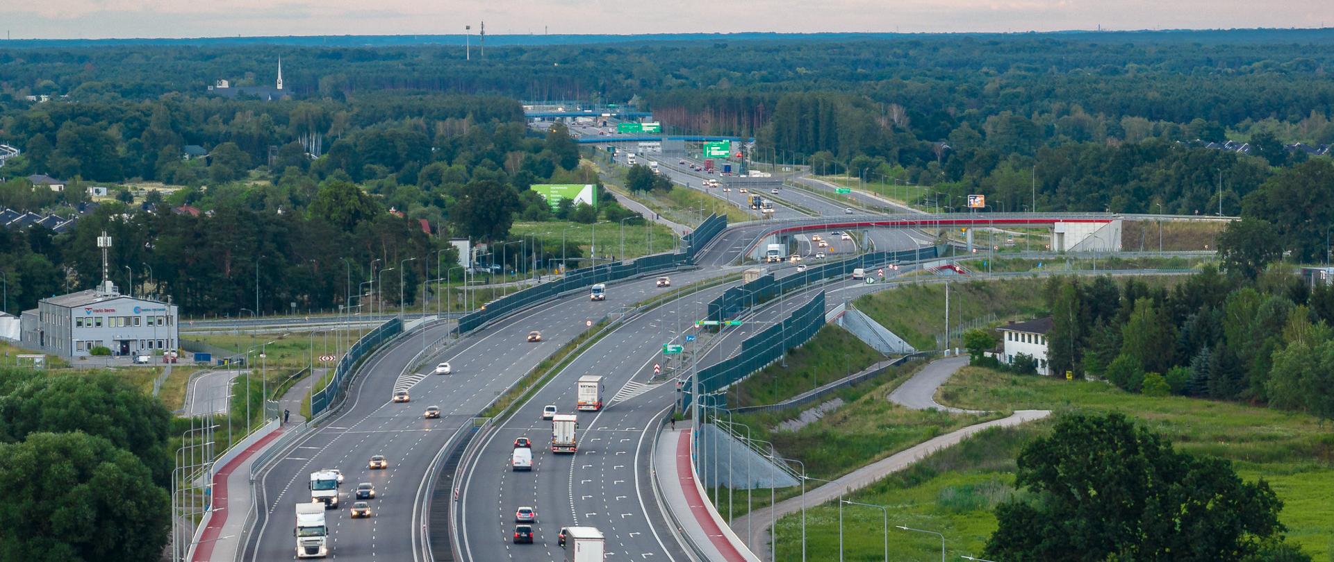 Zdjęcie przedstawia widok z lotu ptaka na drogę ekspresową S2 z wieloma pasami ruchu w obu kierunkach. Na drodze znajduje się kilka samochodów osobowych i ciężarówek. Po lewej stronie widoczna jest ścieżka rowerowa i chodnik, oddzielona od jezdni barierką. W tle widać zielone tereny, drzewa oraz budynki.