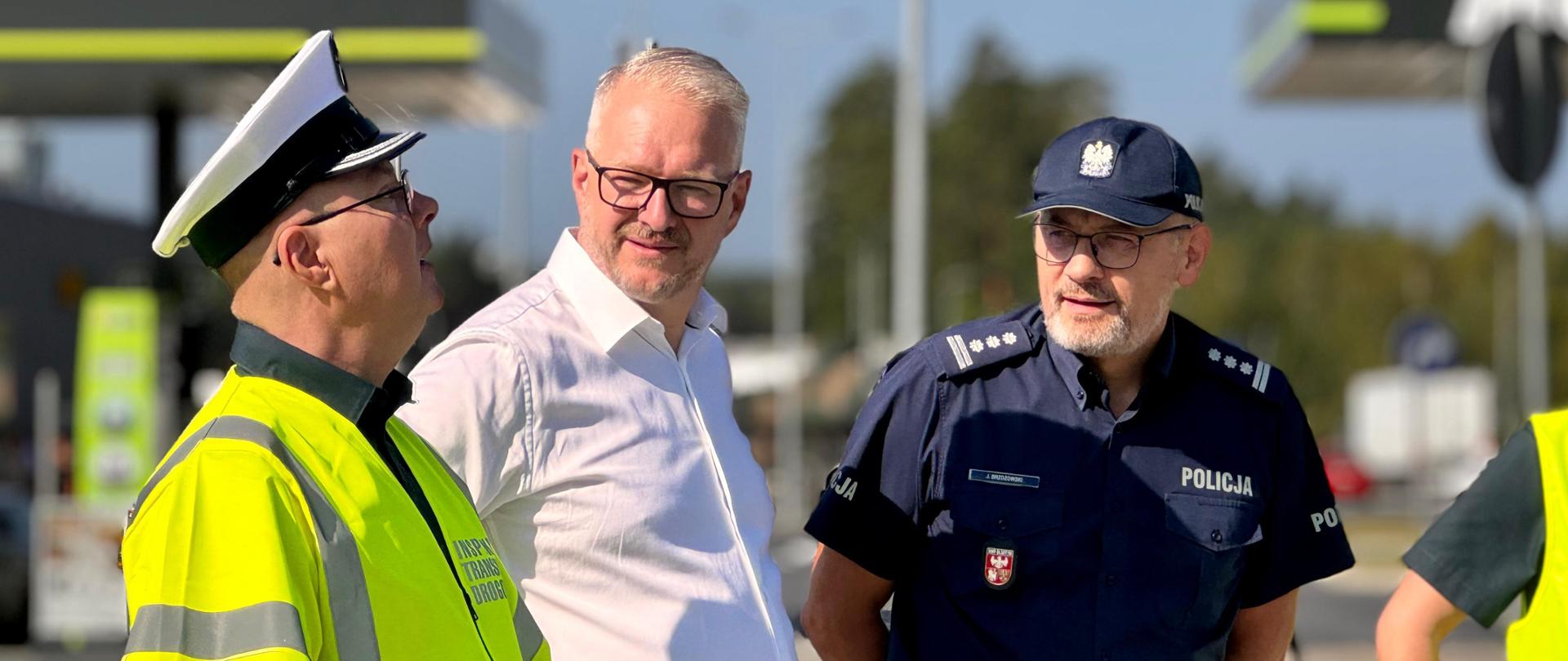 Na zdjęciu trzech mężczyzn, ubrani kolejno w kamizelkę odblaskową, białą koszulę oraz mundur policyjny