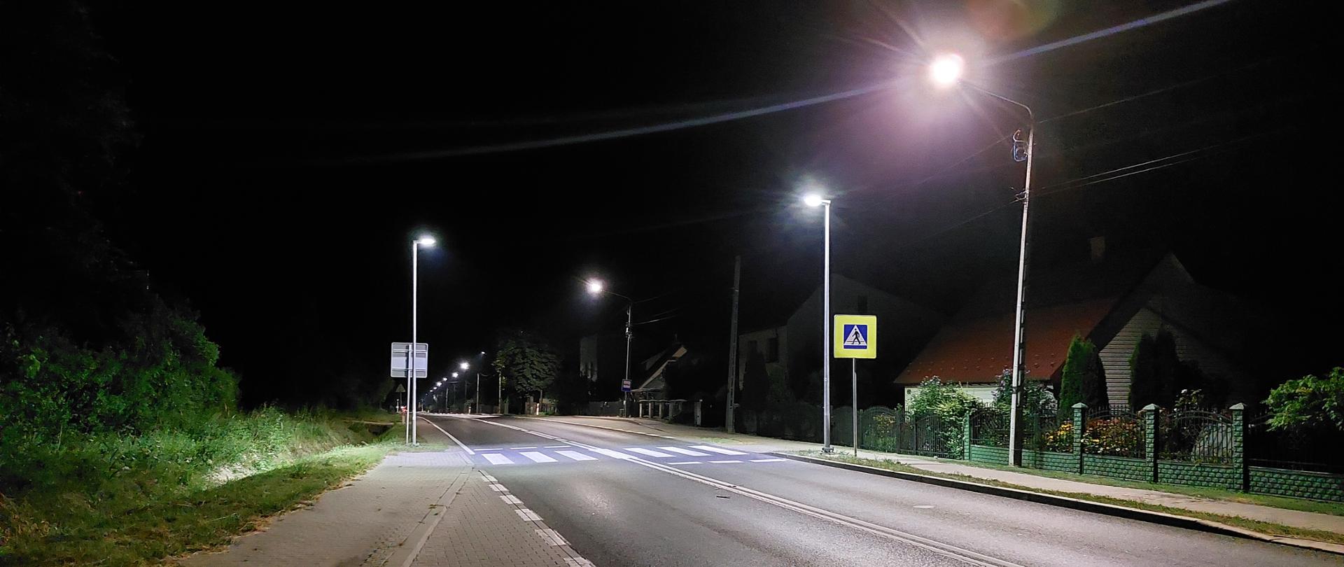 Oświetlone przejście dla pieszych - jednojezdniowa droga nocą, oznakowane przejście dla pieszych, lampy na słupach oświetlające przejście, przy drodze domy