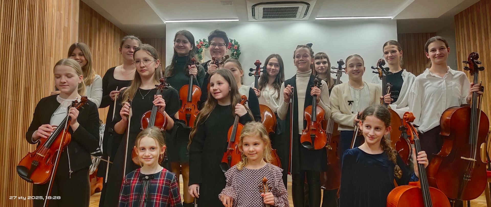 Na zdjęciu widoczna jest grupa młodych muzyków należących do sekcji instrumentów smyczkowych. Uczestnicy są elegancko ubrani, trzymają swoje instrumenty – skrzypce, wiolonczele. Na pierwszym planie znajdują się najmłodsze uczestniczki z radosnym wyrazem twarzy. W tle widoczna jest drewniana ściana oraz świąteczne dekoracje na sali koncertowej. Muzycy są uśmiechnięci i dumni ze swojego występu. Atmosfera zdjęcia jest ciepła i pełna pasji muzycznej.