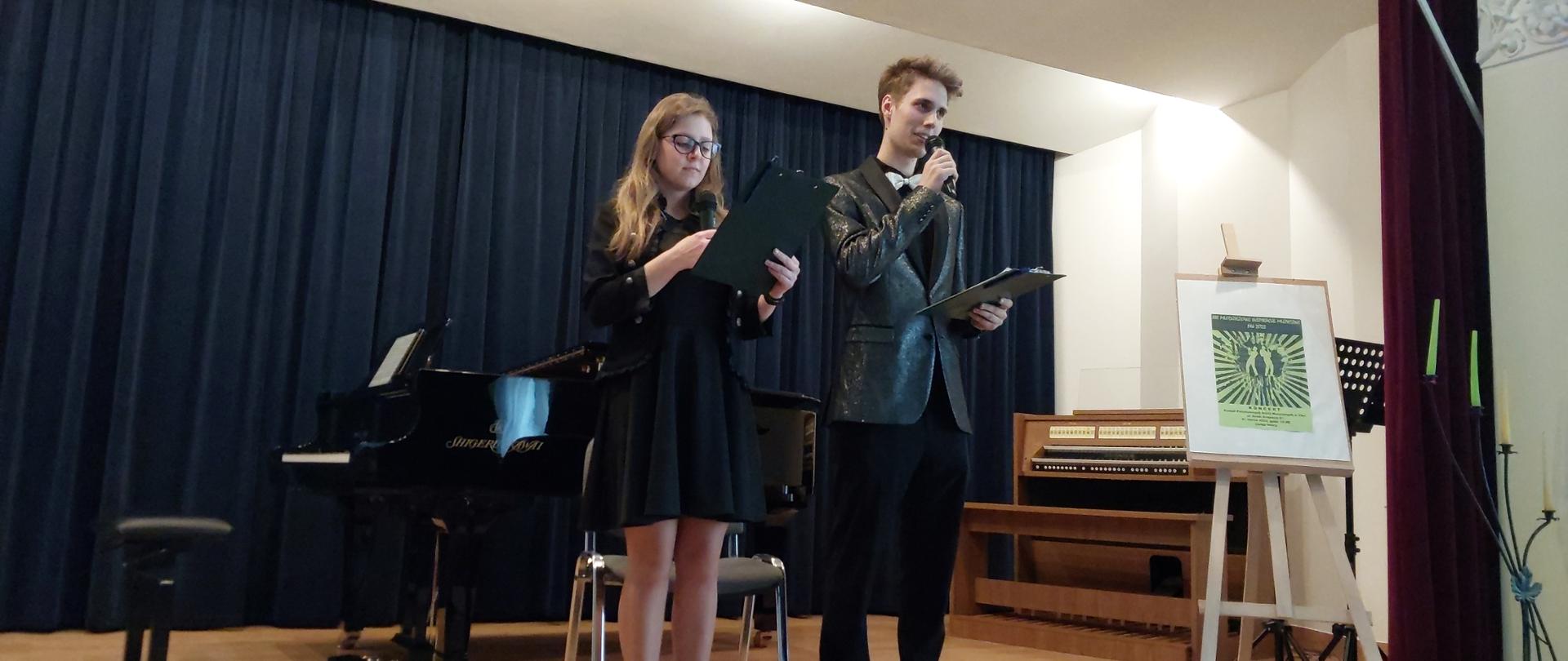 Dwoje uczniów stoi na scenie i zapowiada koncert. W tle fortepian i elektroniczne organy. 