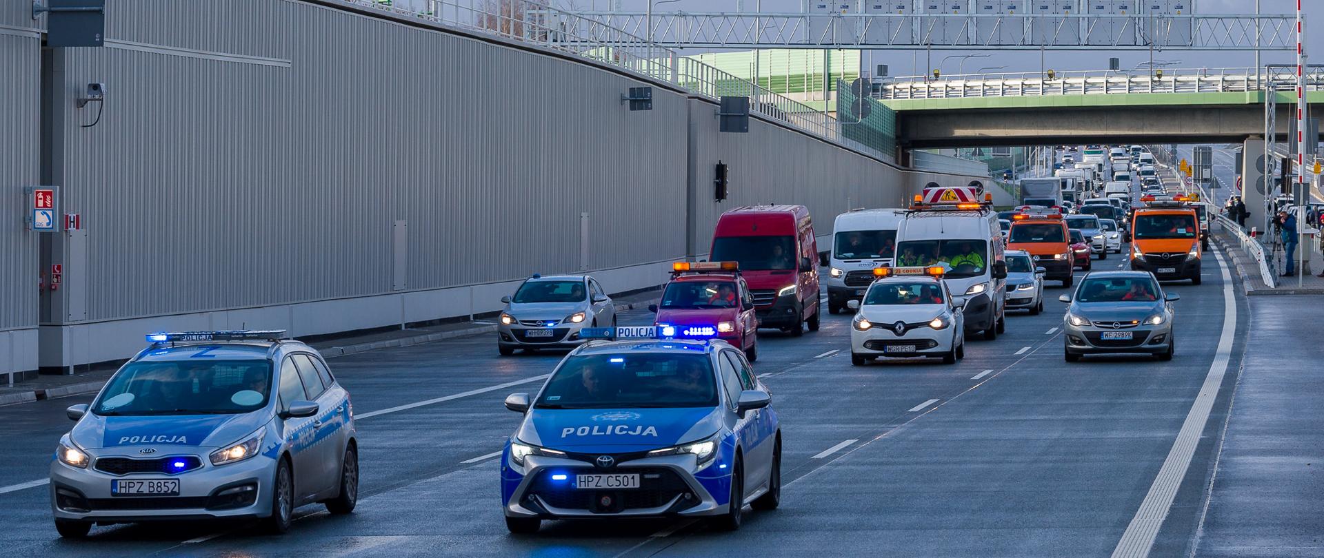 Zdjęcie przedstawia scenę na wielopasmowej drodze z wieloma pojazdami, w tym dwoma radiowozami z migającymi niebieskimi światłami, czerwonym vanem oraz kilkoma innymi samochodami i pojazdami serwisowymi GDDKiA. Wszystkie pojazdy poruszają się za dwoma radiowozami na pierwszym planie.