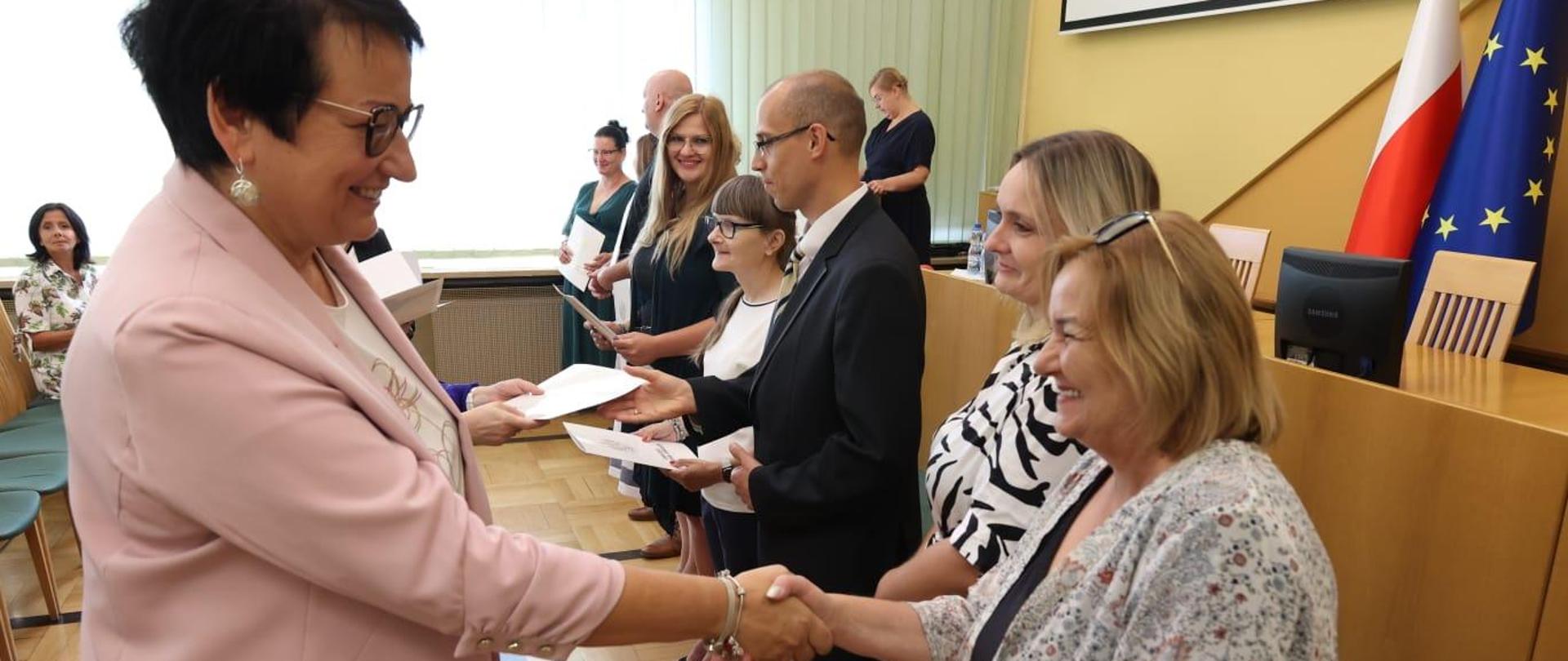 Wojewoda Opolski wręcza jednej z nauczycielek akt nadania stopnia awansu. Ściska jej dłoń w ramach gratulacji. Obie się uśmiechają. W tle widać pozostałych awansowanych, którzy już otrzymali swoje akty. 