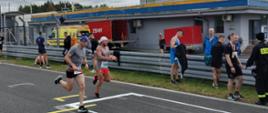 Dwóch zawodników wbiega na start trzymając pałeczki obok stoją inni uczestnicy biegu niedaleko stoi karetka pogotowia.