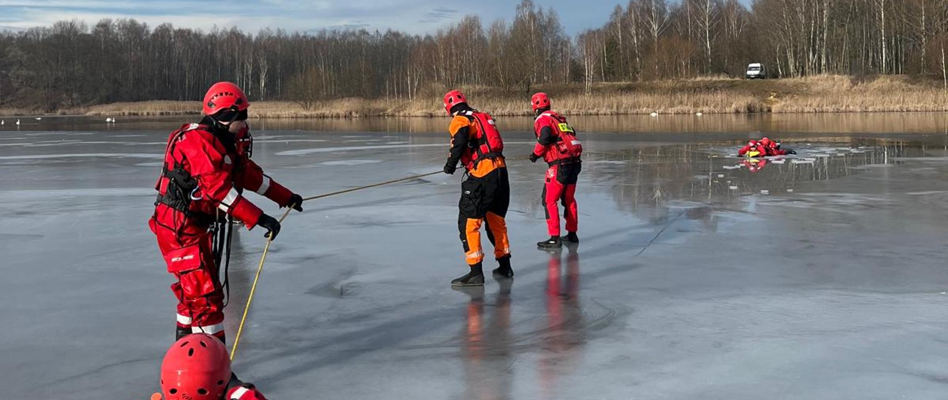 Część praktyczna warsztatów lodowych dla dowódców zmian funkcjonariusze ubrani w suche skafandry oraz kamizelki asekuracyjne i kaski ćwiczą akcję ratowniczą za pomocą pasa węgorza