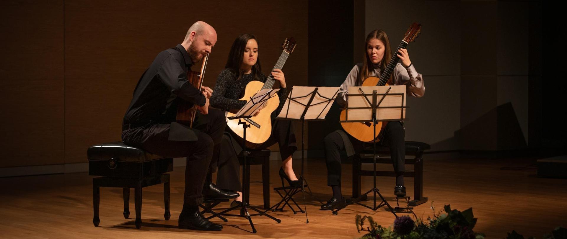 Zdjęcie przedstawiające grających (na gitarach klasycznych) gitarzystę oraz dwie gitarzystki. Zdjęcie zrobione w czasie koncertu na sali koncertowej szkoły muzycznej, przed artystami pulpity z nutami, przed nimi podnóżki gitarowe.