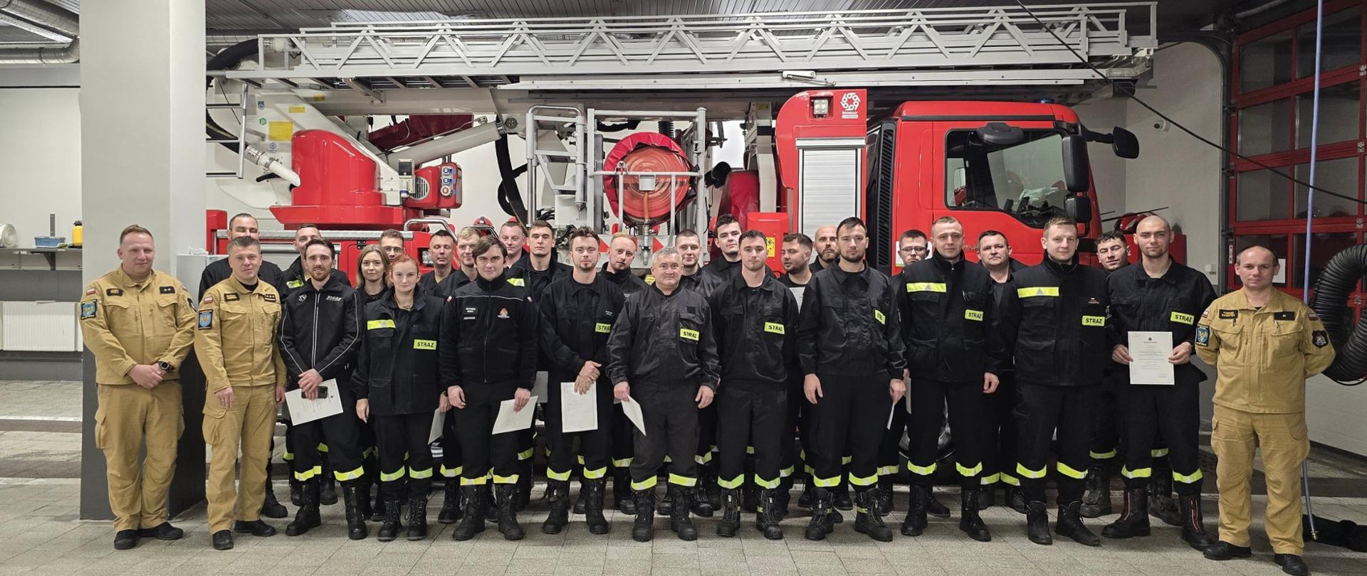 Na zdjęciu znajdują się strażacy w koszarówkach ustawieni do zdjęcia w garażu na tle podnośnika