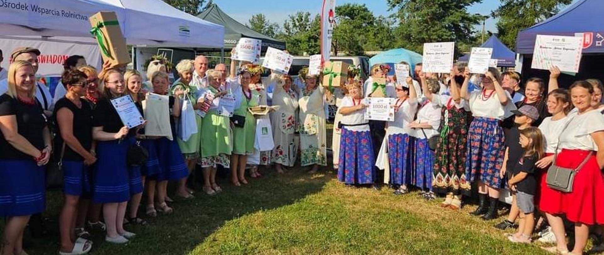 Duża grupa osób, w tym większość kobiet w strojach regionalnych, stojąca na tle namiotów ze stoiskami informacyjnymi różnych instytucji i firm.