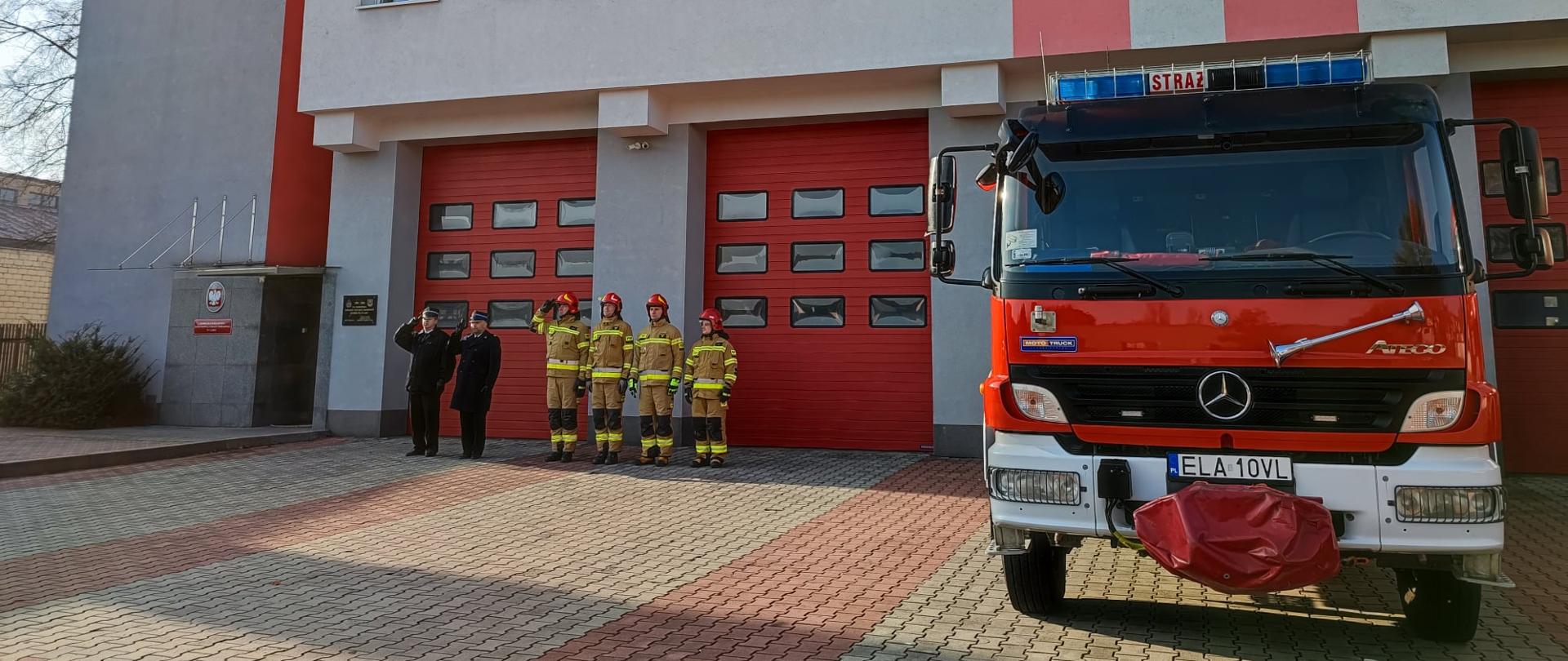 Zdjęcie przedstawia strażaków Komendy Powiatowej Państwowej Straży Pożarnej w Łasku, podczas podniesienia flagi państwowej oraz uroczystego odśpiewania hymnu narodowego.