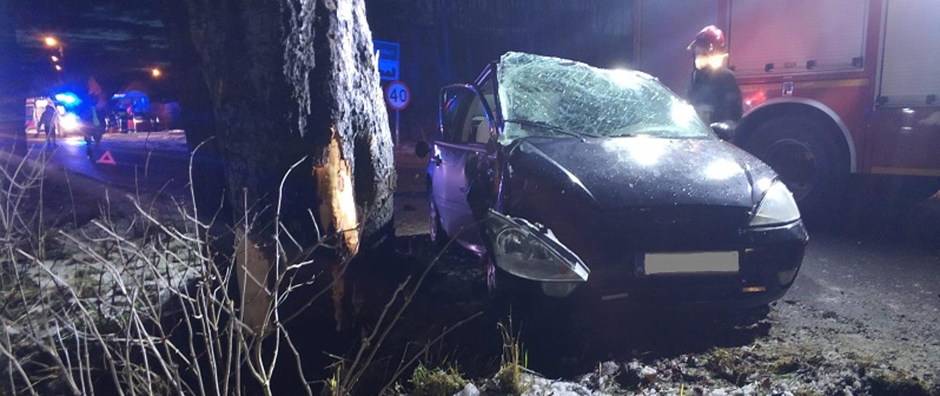 Zdjęcie wykonane nocą, na pierwszym planie widoczny przodem samochód osobowy Ford Focus koloru granatowego. Rozbita szyba przednia i zwisający nad kołem reflektorem. Z lewej strony pojazdu widoczny fragment drzewa z uszkodzoną korą po uderzeniu pojazdu.
