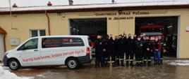 Zdjęcie grupowe strażaków ochotniczej straży pożarnej na tle remizy Ochotniczej Straży Pożarnej w Jelonkach. Po lewej stronie stoi samochód z napisem „Łączy nas krew, która ratuje życie”