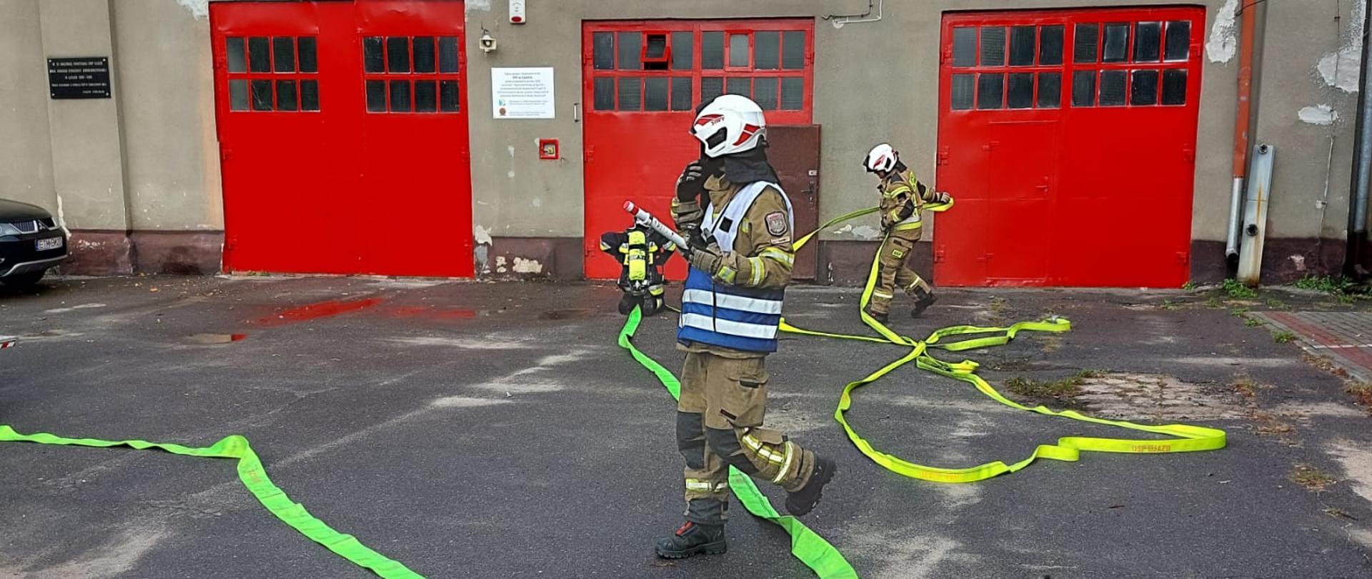Przed budynkiem strażnicy strażacy rozwijają na asfaltowym podłożu jasno zielone węże pożarnicze.
