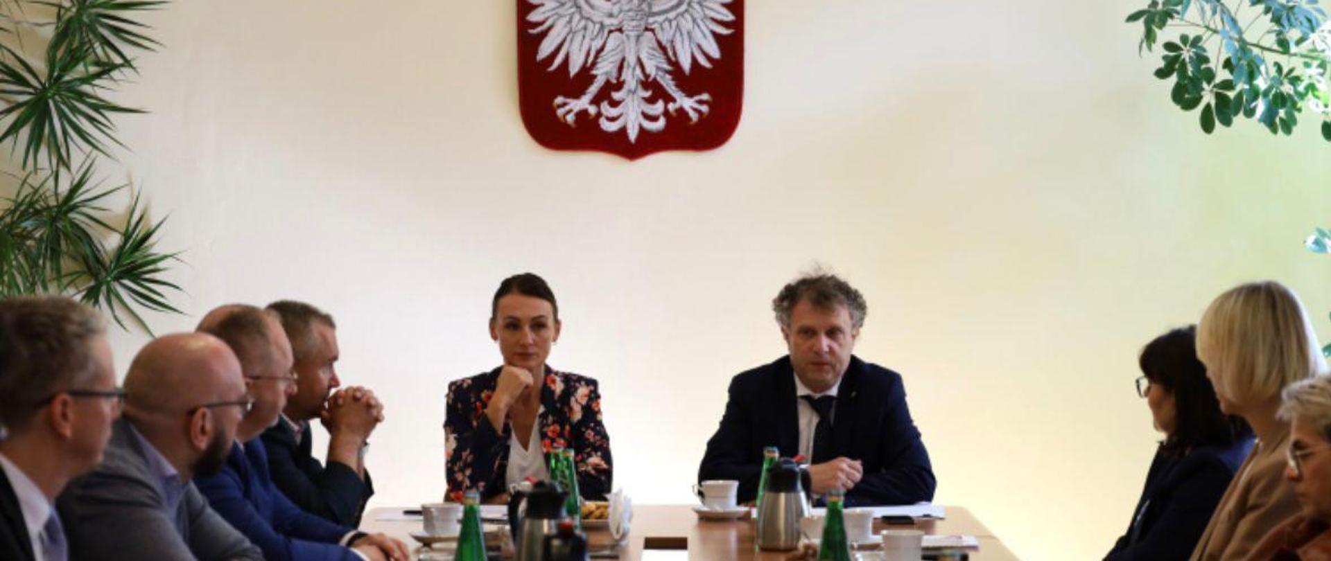 Dwie osoby siedzą obok siebie w oddali przy stole. Pierwszy od lewej siedzi wiceminister Jacek Karnowski.
