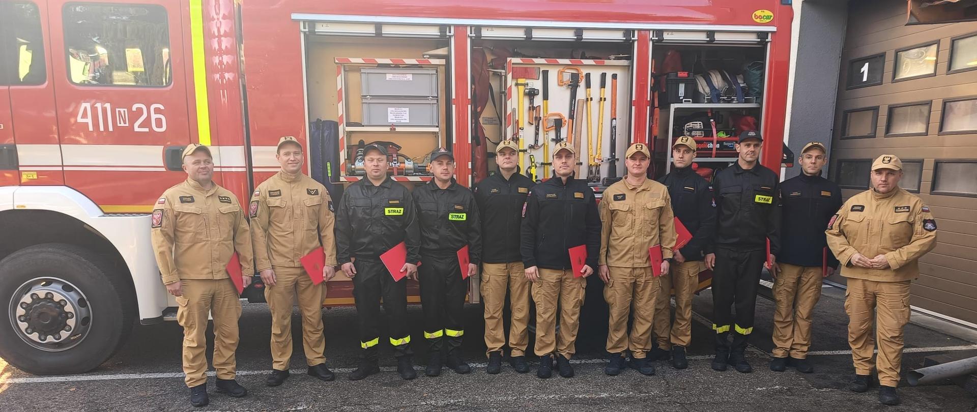 grupa strażaków w mundurach ustawionych w jednym szeregu na tle dużego czerwonego samochodu strażackiego