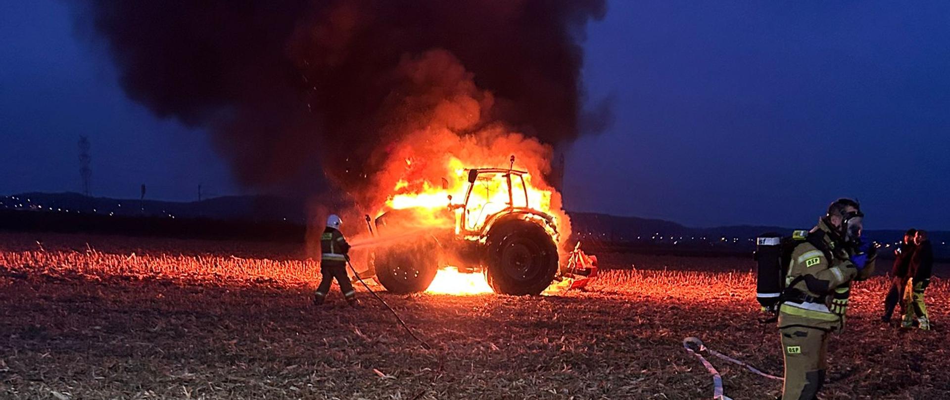 Na zdjęciu znajdują się strażacy, którzy gaszą palący się ciągnik rolniczy na polu. 