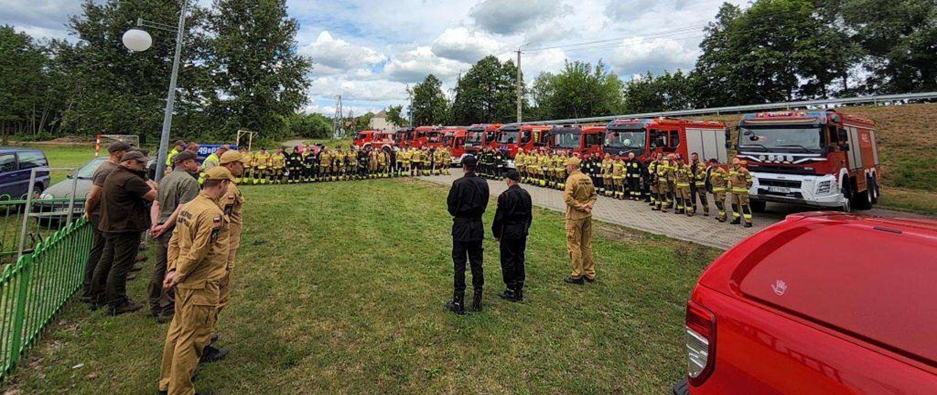Strażacy stoją na tle pojazdów pożarniczych