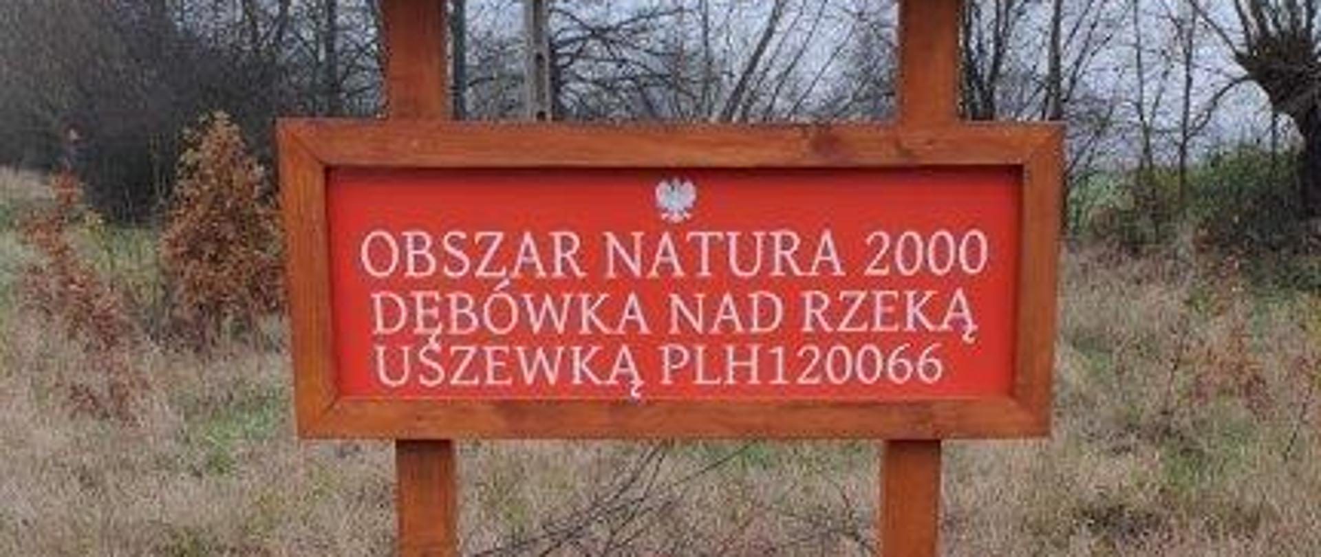 Drewniana tablica z daszkiem. Na czerwonym tle znajduje się biały napis - Obszar Natura 2000 Dębówka nad rzeką Uszewką PLH120066