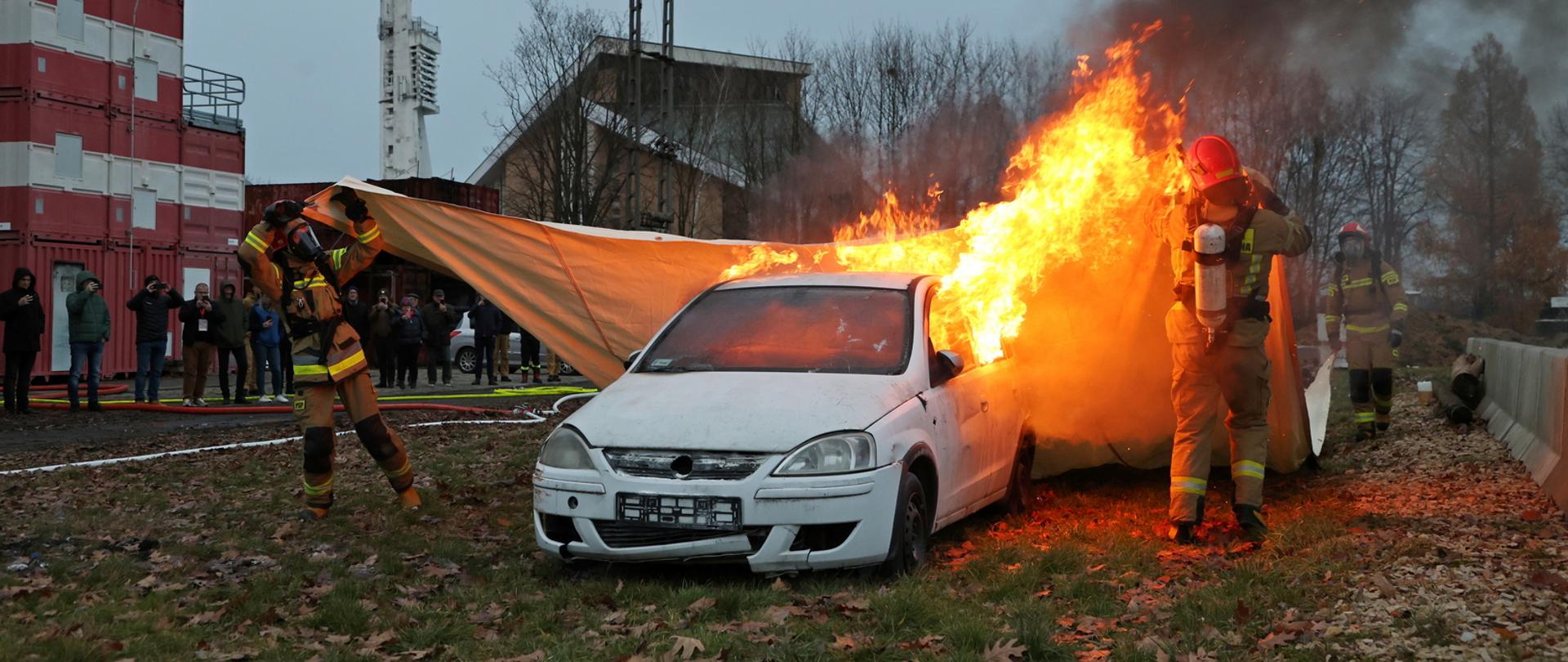 Poligon szkolny, dwóch strażaków w ubraniach specjalistycznych z maską rozciąga płachtę gaśniczą nad płonącym wrakiem samochodu, w głębi stojący obserwatorzy pokazu