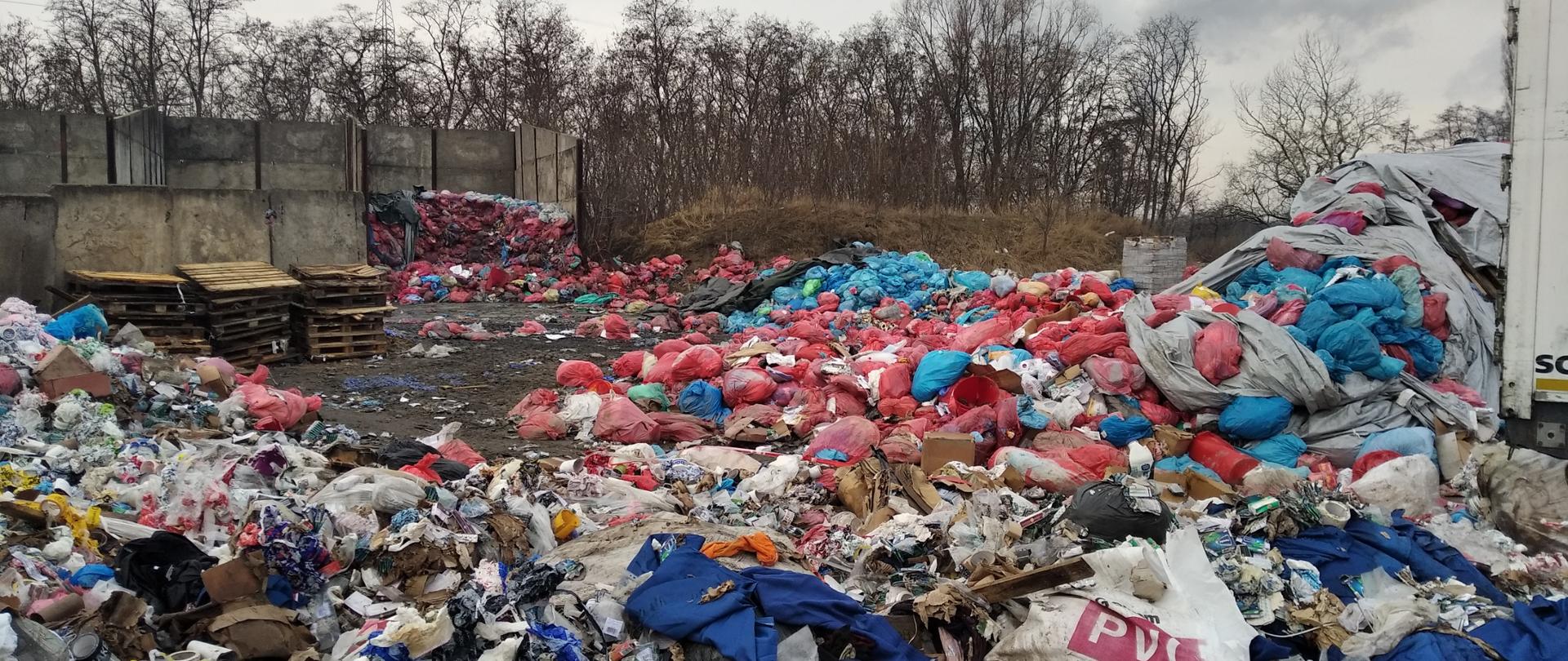 Setki toreb plastikowych z odpadami zalegają pod gołym niebem. Część z nich jest porozrywana. W tle widać betonowy płot i stojące za nim drzewa.