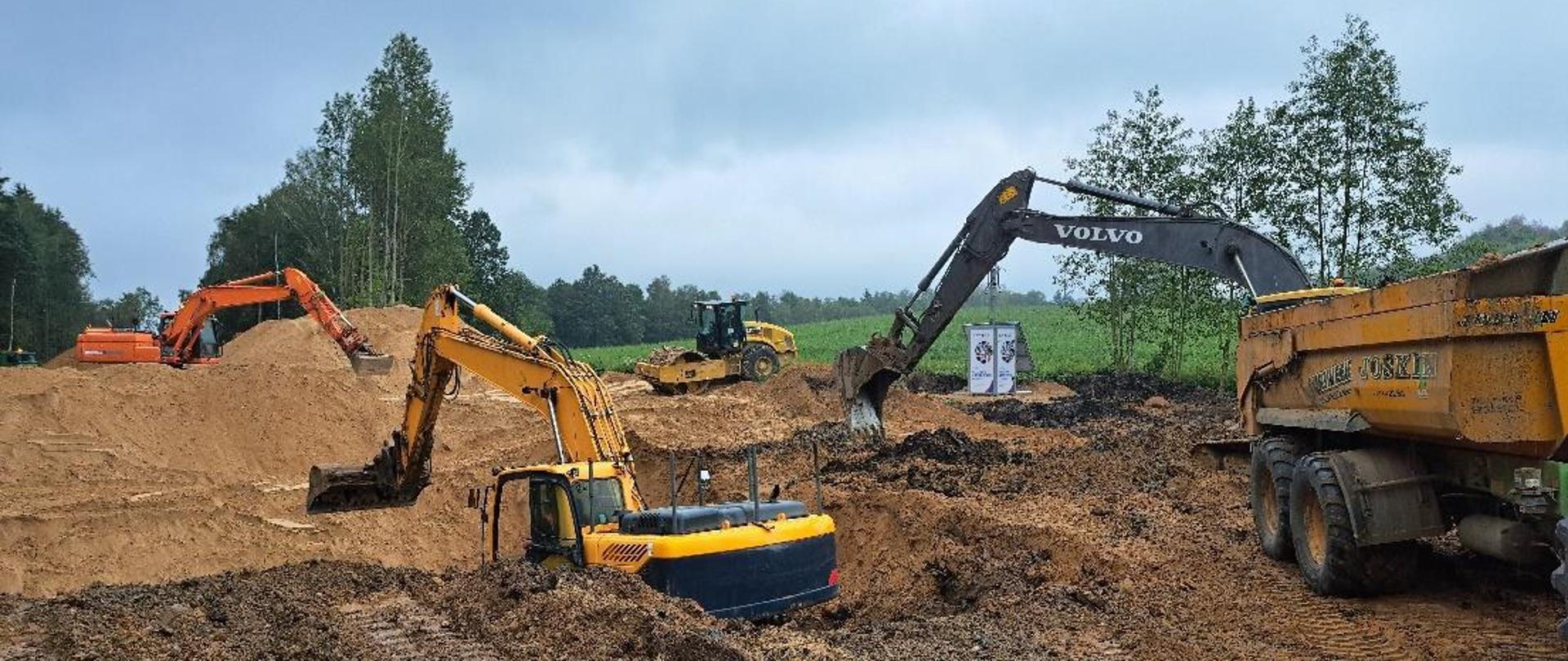 Na zdjęciu widoczne prace ziemne przy użyciu trzech koparek i jednego walca., widać nasyp ziemny. W oddali zielone pole i linia drzew.