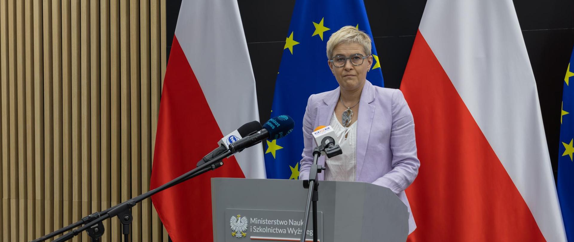 Wiceminister Mrówczyńska stoi za mównicą, za nią pod ścianą flagi Polski i UE.