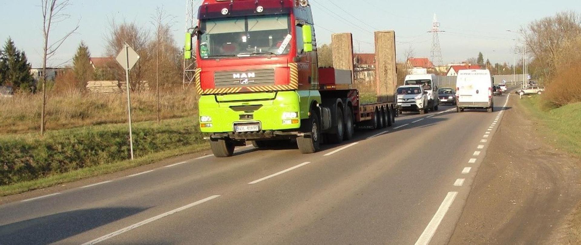 Na pierwszym planie ciężarówka poruszająca się jednojezdniową drogą krajową. W tle zabudowania i ekrany akustyczne.