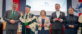 Minister Wieczorek stoi w szeregu obok mężczyzny w zielonym ceremonialnym stroju z gronostajowym kołnierzem, dwóch kobiet i mężczyzny w garniturze, za nimi na ścianie wielki ekran z napisem na białym tle Uniwersytet Radomski.