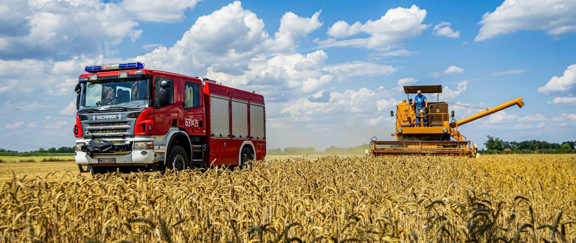 Na zdjęciu rolnik podczas zbioru ziarna na polu oraz w tle czerwony samochód Straży Pożarnej.
