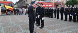 Trzech strażaków na środku placu apelowego otrzymuje od Komendanta awanse. Za nimi w rzędzie stoją inni strażacy, w Tle dwa samochody pożarnicze i budynki.