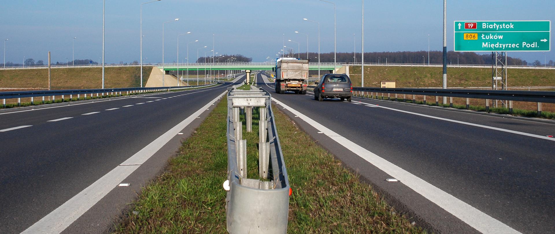 DK19 obwodnica Międzyrzeca Podlaskiego, odcinek dwujezdniowy w obrębie węzła Międzyrzec Podlaski Południe. Widok na dwie jezdnie z poziomu drogi, centralnie pas rozdziału z barierą energochłonną. Na jedni po prawej jadą pojazdy, na dalszym planie wiadukt i znaki drogowe 