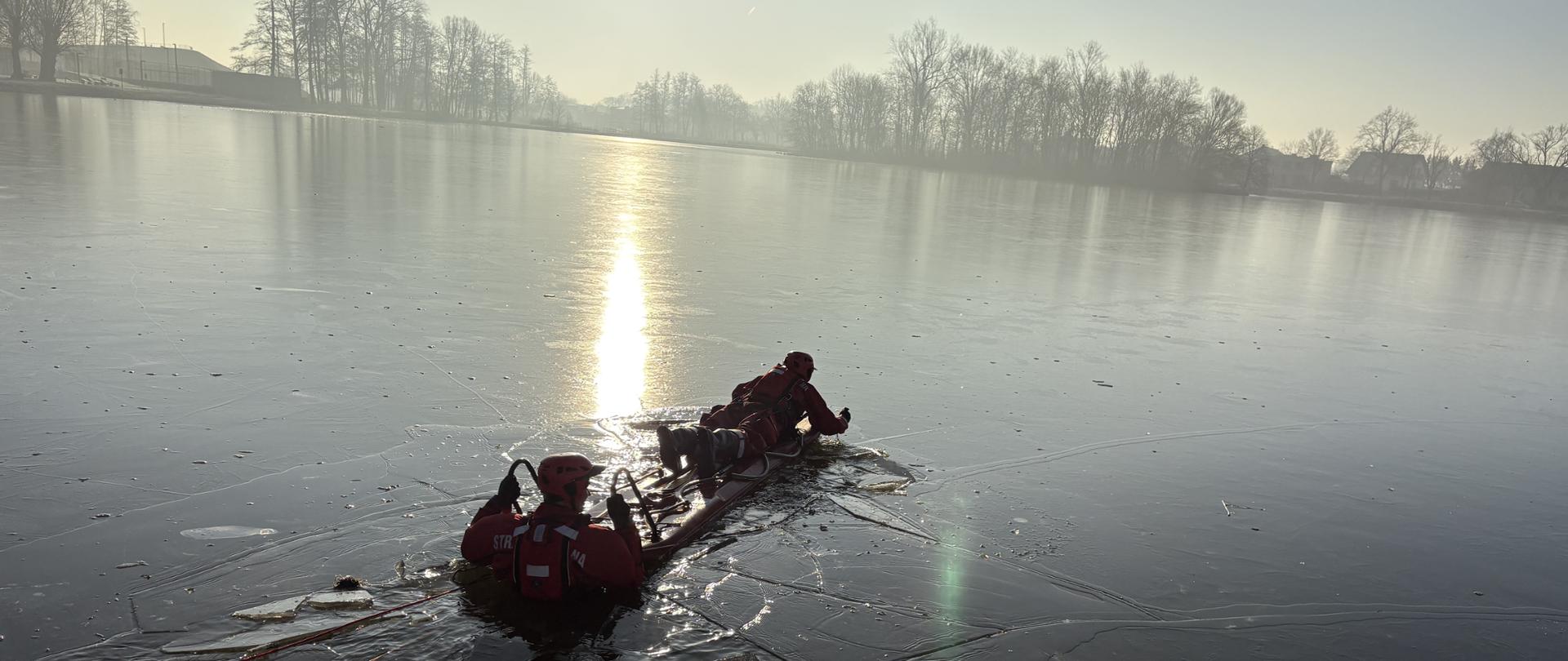 Strażacy ćwiczą poruszanie się po lodzie za pomocą sań lodowych. Ćwiczenia odbywają się na akwenie z cienką pokrywą lodu, który kruszy się pod ratownikami