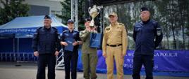 Funkcjonariusze Policji i Państwowej Straży Pożarnej stoją obok ucznia zwycięzcy konkursu. Uczeń trzyma puchar i nagrody.