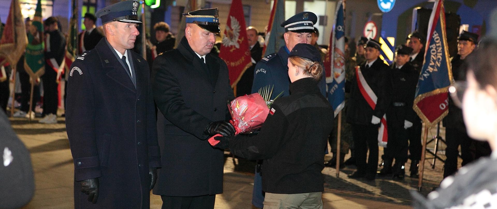 Plac przed Pomnikiem Żołnierza Polskiego w Rawiczu. Delegacja rawickich służb mundurowych składa wiązankę kwiatów. W tle poczty sztandarowe uczestniczące w uroczystości.