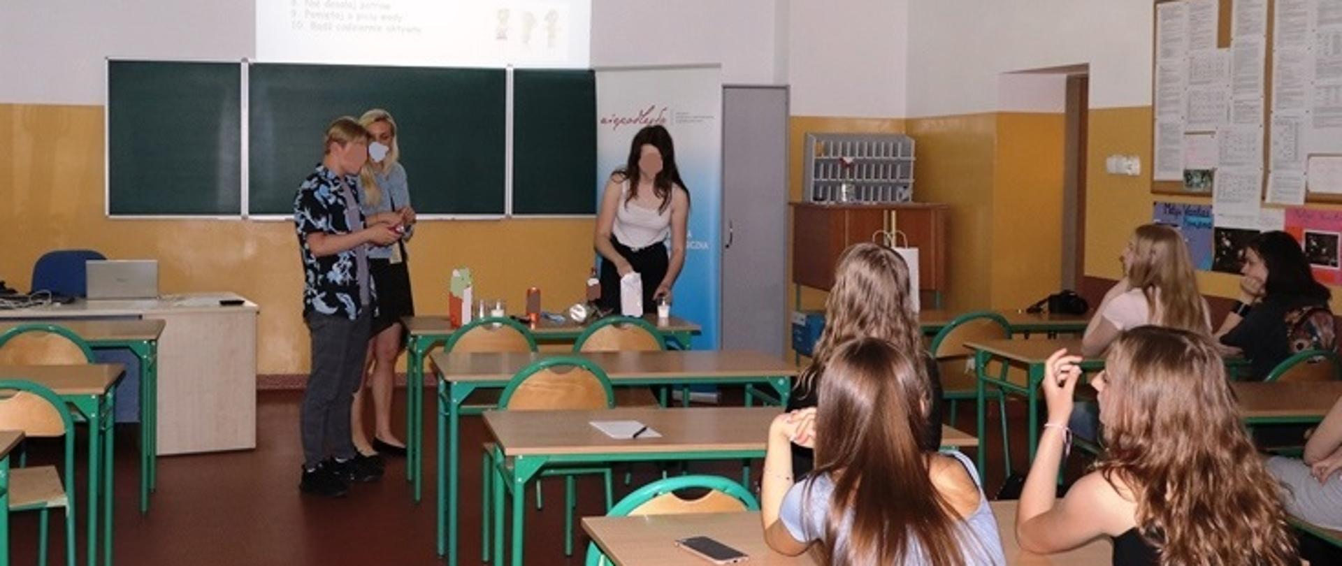 Zdjęcie przedstawia pracownika WSSE w Gorzowie Wlkp., ucznia i uczennice liceum w trakcie warsztatów (sprawdzanie ilości cukru w popularnych produktach spożywczych). Po prawej stronie zdjęcia widoczni są inni uczniowie siedzący w ławkach.