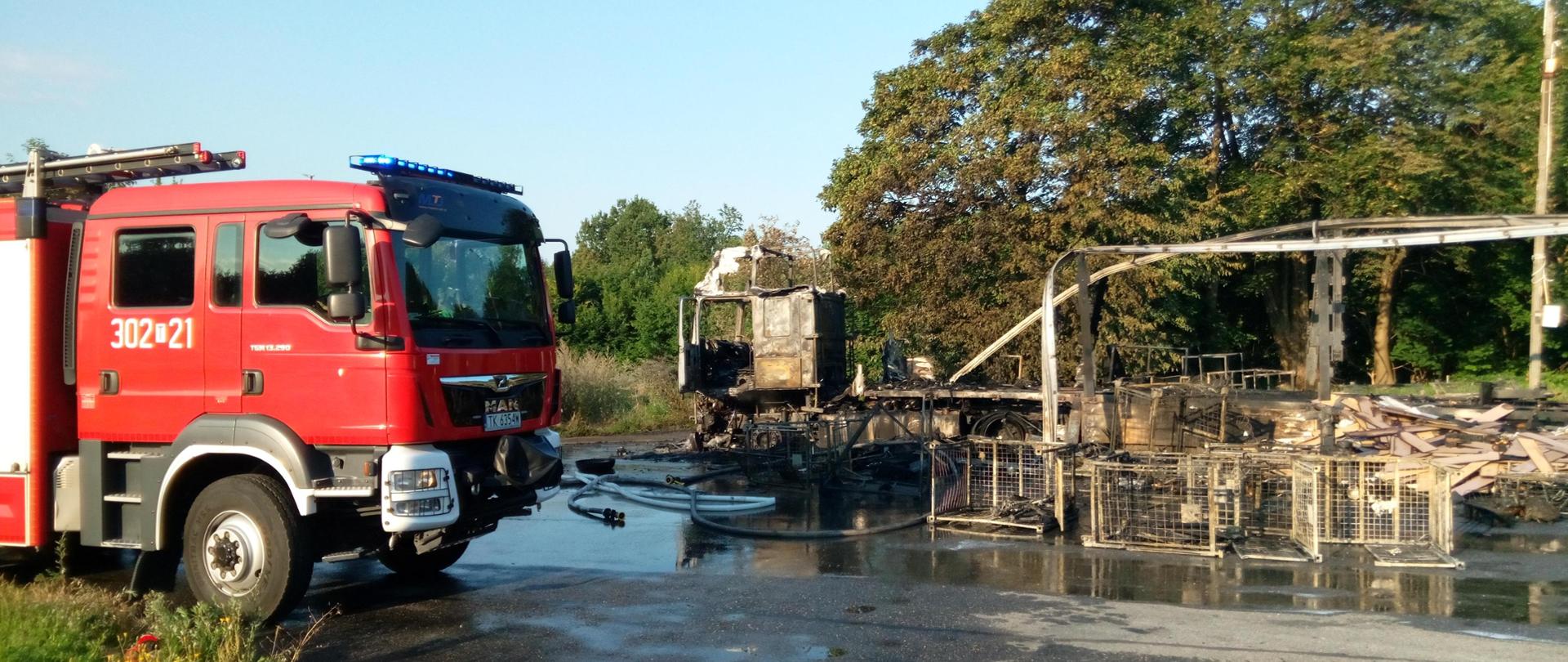 Zdjęcie przedstawia spalony zestaw ciągnika siodłowego i naczepy. Po lewej stronie samochód pożarniczy.