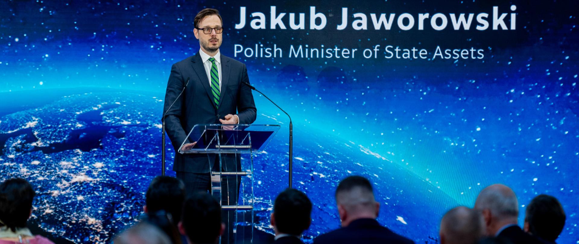 Minister Jakub Jaworowski stoi za mównicą. W tle ekran, na którym widać kulę ziemską. Ujęcie zza słuchaczy. 